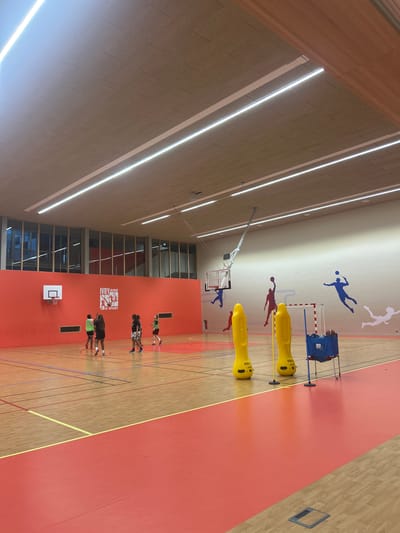 A brightly lit indoor gymnasium in Ivry-sur-Seine, France is photographed from an elevated perspective. Several people are present on the wooden floor, which is marked with various colored lines and sections of solid red. The people appear to be engaged in sports activities. Various pieces of sports equipment, including yellow cylindrical objects, a net with blue equipment behind it, and basketball hoops, are located throughout the space. Artwork depicting athletes is affixed to the upper walls. Large windows are visible, as well as rows of overhead lights. A logo of "Ivry aime le Sport" is visible on the wall.
