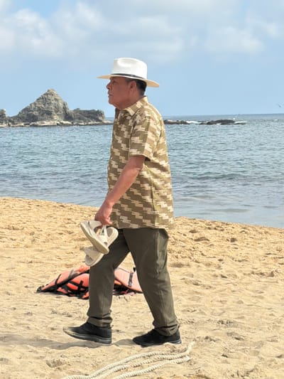 An elderly Asian man stands on a sandy beach near the ocean. He wears a white hat, patterned shirt, olive pants, and black shoes. He holds a pair of light-colored sandals in his hand. Behind him, the ocean stretches out with rock formations visible in the distance. A life vest and coiled rope are on the sand beside him. The sky is light blue with scattered clouds.