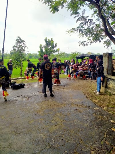 The image shows a group of people gathered in an open area in Kecamatan Kedawung, Indonesia. The group is comprised of 20 individuals, standing in a semi-circle around a truck, with some seated and others standing. Some people are wearing traditional clothing with yellow and red accents, while others are dressed in casual attire. Several people are interacting with each other while others are focused on the action happening in front of them. The scene is set in a rural environment with a grassy field and a dirt path leading to the group.  There are several black plastic buckets scattered on the ground in front of the group. The trees are bare and the sky is overcast with a few wispy clouds. The overall feel of the image is calm and peaceful. The scene is a celebration of some kind, perhaps a cultural event or a religious ceremony.  The image is a snapshot of a moment in time, and it captures the essence of community and tradition.