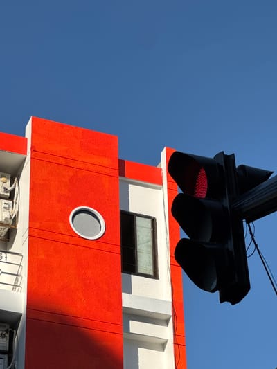 A traffic light with a lit red signal is positioned in the foreground against a bright blue sky. To the left of the traffic light, a section of a building with a striking orange facade is visible. The building has white trim and features a circular, grey-tinted window. The lower part of the orange facade is partially obscured by a shadow. The scene suggests an outdoor urban setting.