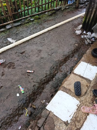 A wet, paved path with scattered litter runs beside a white PVC pipe along a wooden fence. Rainwater flows along the path's edge, near a tiled area. Here are assorted sandals and a small tire. The scene conveys a sense of neglect and disrepair.