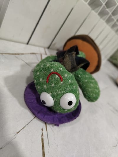 A green plush toy with white, googly eyes, a red smile, and a purple felt base sits on a white, patterned surface. It wears a brown hat with a black brim, reminiscent of a mushroom. The background includes a white, slated structure. The toy's textured green fabric suggests a handmade quality.