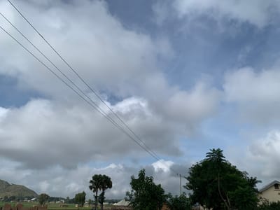 This image captures a wide-angle view of a cloudy sky over Zawan, Nigeria. Three parallel black wires traverse the upper portion of the frame, stretching diagonally from the upper left towards the right. Below the wires, a landscape unfolds with rolling hills in the background on the left, dotted with small buildings. In the middle ground, a scattering of homes and structures are visible amidst green fields. Several trees are present, including a distinctive palm tree with its fronds fanned out on the left, and other leafy trees. The foreground shows the partially constructed brick walls of a building. The sky is filled with a mix of white and gray clouds, with patches of blue sky visible in the upper right. The overall impression is of a semi-rural setting under an overcast sky.