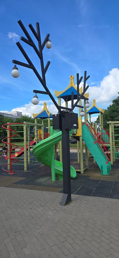 A children's playground is depicted in a city setting, likely in Bryansk, Russia, under a partly cloudy blue sky. The foreground features a paved walkway made of grey rectangular bricks. Behind the walkway is the playground, which includes a black, tree-like lamppost with several white globe lights. The playground equipment is colorful and constructed from various materials. Prominently visible are a bright green spiral slide and a green straight slide with red railings. Other elements of the playground include climbing structures with yellow and blue canopies, red spiral climbing bars, and green netting. The ground surface of the playground is made of black rubber mats. In the background, there are green trees and a multi-story building. Two individuals, a woman and a child, are partially visible on the left side of the playground, near the red spiral structure. The scene captures a typical urban playground environment.