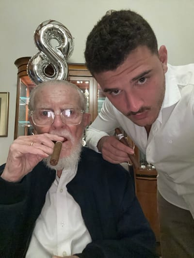Two men pose for a selfie while holding cigars. The younger man is behind and above the older man, both facing the camera. A silver "80" balloon floats near the older man's head. They're dressed in collared shirts. The older man wears glasses and a dark jacket. A wooden cabinet with glassware is in the background.
