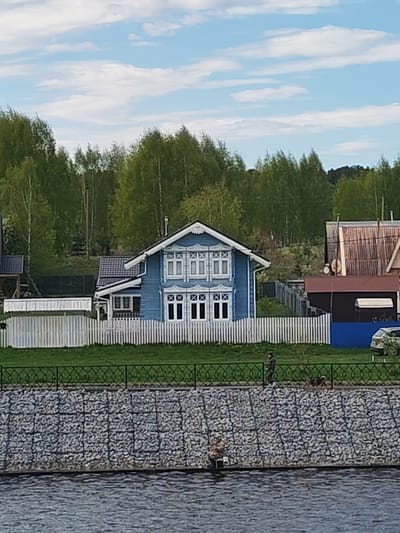 The image depicts a blue, two-story wooden house with white trim and a white picket fence in Myshkin, Russia. The house is set against a backdrop of green trees under a partly cloudy sky. To the right of the blue house, a brown and red-roofed building is visible. In the foreground, there is a stone embankment with a decorative black fence. A person walking along the embankment is visible behind the fence. The water level suggests the scene is near a river or lake. A light-colored car is parked near the house on the right.