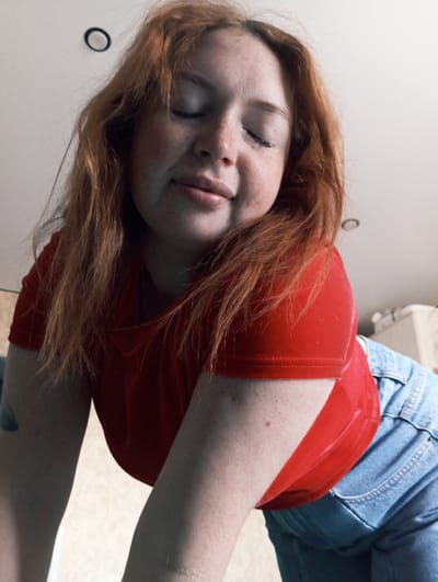 A fair-skinned, freckled person with red hair is seen lying down. The person is wearing a red shirt and blue jeans. The background appears to be a plain white wall and a light-colored floor. There is a small circular object on the wall. The location is not identifiable as Yurgamysh, Russia from the visual data provided.