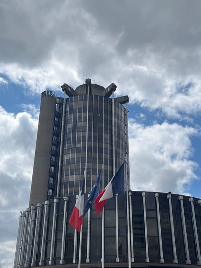 This is a medium shot, taken from a low angle, of a modern, multi-story building. The building features a dark glass facade with horizontal white accents. The top of the building has a unique, angular design with protruding architectural elements. Two flags are prominently displayed, waving in the wind. One flag is the French tricolor, and the other appears to be the European Union flag, with its circle of yellow stars on a blue background.
The sky is filled with a mix of dramatic, grey storm clouds and patches of bright blue sky, suggesting a dynamic weather pattern. The lighting indicates it is daytime, likely midday or early afternoon, given the brightness of the clouds and blue sky. The architectural style of the building and the presence of the French and EU flags strongly suggest a location in France, specifically hinted at as Créteil.
There is no discernible activity or event taking place other than the flags waving and the clouds moving. The scene evokes a sense of urban architecture and national or supranational identity, set against a backdrop of changing weather. No visible text is present in the image.