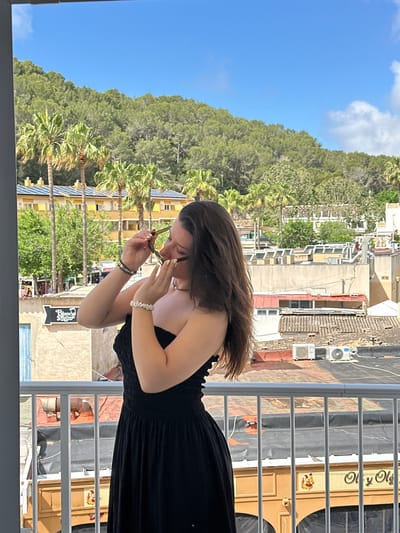 The image shows a woman standing on a balcony, holding sunglasses in her hands. She is wearing a black dress and has a white bracelet on her wrist. In the background, there is a view of buildings, palm trees, and a forested hill under a blue sky. The balcony has a white metal railing. The location is Calvià, Spain.