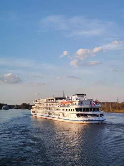The image shows a white passenger cruise ship, named "СЕРГЕЙ ДЯГИЛЕВ", sailing on a river. The ship has multiple decks and a Russian flag is visible at the stern. Another smaller white ship is further down the river, towards a bridge or land structure. The water is dark blue with ripples. The riverbanks are lined with trees, mostly bare, indicating a cooler season. Power lines are visible in the background. The sky is blue with scattered white clouds. The scene likely takes place in or near Базарово, Russia, given the name on the ship.