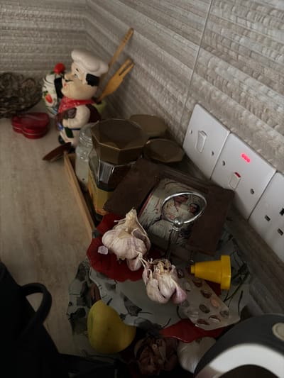 The image shows a collection of objects on a countertop, likely in a kitchen, in Dromolaxia - Meneou Municipality, Cyprus. In the foreground, there are several bulbs of garlic, a yellow object that appears to be a funnel or stopper, a patterned dish with what looks like scattered seeds or beans, and a piece of red ceramic or plastic. Behind these, a small framed image, possibly a religious icon, is propped up. A magnifying glass with a silver handle rests on top of the frame. Further back, a glass jar filled with coffee grounds is visible, topped with a hexagonal lid. To the left, a ceramic figurine of a chef with a red bandana and holding kitchen utensils stands near a heart-shaped red container and a decorative metal bowl. The background features a tiled wall with a light beige, textured pattern. Electrical outlets and light switches are mounted on the tiled wall on the right side of the image. The lighting suggests it is daytime.