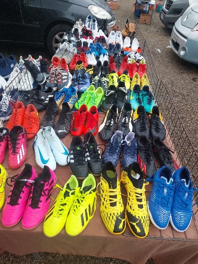 The image presents an outdoor market stall in Eckbolsheim, France, showcasing a large collection of athletic footwear. Hundreds of pairs of shoes, primarily soccer cleats and indoor soccer shoes, are displayed across a brown, fabric-draped surface. Some footwear is organized on metal wire grids. The shoes exhibit a wide palette of colors, including neon yellow, bright pink, red, blue, black, white, and various multi-color designs, featuring visible branding elements such as Adidas stripes, Nike swooshes, and Puma logos. The background consists of an asphalt or gravel surface with multiple parked vehicles: a dark-colored minivan or hatchback on the left, and a silver/grey car partially visible on the right. Another dark-colored vehicle is present further in the background. No individuals are visible in the frame interacting with the merchandise.