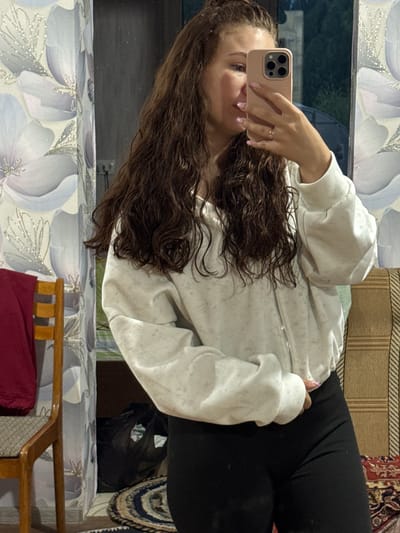 A young woman with long, brown, wavy hair is taking a selfie using a pink smartphone. She is wearing a light gray zippered sweatshirt and black leggings. She is wearing a gold ring. The background includes a floral pattern on what appears to be a bedspread or mattress, part of a wooden chair frame with red fabric, and a rug with various patterns. The location is not identifiable as the city of Nizhnyaya Gar', Russia from the given image.