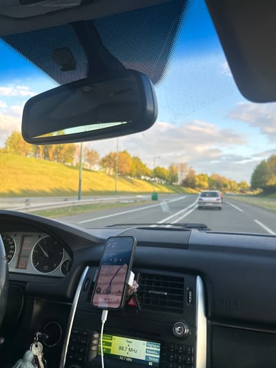The image shows the interior of a car, likely a Mercedes, with the driver's perspective facing the road. The car is in motion on a multi-lane highway in what appears to be a rural area. Through the windshield, one can see a blue sky with scattered clouds, green fields, and trees lining the side of the road. A car is visible ahead in the same lane.

Inside the car, a smartphone is mounted on the dashboard and displaying navigation with an estimated arrival time of 20:19. The dashboard features a radio display tuned to 88.7 MHz. The car's speedometer is partially visible and reads 635.0km. Keys are visible hanging from the dash. The location appears to be near Lyon, France.