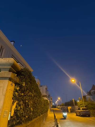 The image shows a street scene in Cascais, Portugal, at dusk or early evening. The sky is a deep blue, with a hint of a faint contrail or atmospheric phenomenon visible in the upper left quadrant, and a brighter, streaking light source diagonally bisecting the sky from the upper right to the center. On the left, a building with a white facade and a dark-capped chimney is partially visible, with a substantial hedge or ivy-covered wall running along its base. A yellow post with signage, including a triangular warning symbol, is positioned in front of the hedge.
The street itself is paved with cobblestones and slopes downwards from right to left. Several streetlights illuminate the scene, with one prominent streetlight in the mid-right casting a bright yellow glow and a long, diagonal beam across the sky. A white scooter with its headlights on is moving down the street towards the viewer. Further down the street, several cars are parked or moving, and more streetlights are visible. The overall atmosphere is tranquil and residential, characteristic of a European coastal town in the evening.