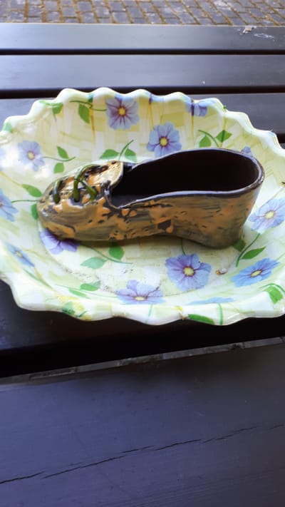 A ceramic novelty shoe, likely a planter or decorative item, sits on a decorative, scalloped-edge plate with a floral pattern. The shoe is dark brown with an orange glaze streaked across its surface. Small green sprouts are visible emerging from the front of the shoe. The plate has a light green background with repeating blue flowers with yellow centers and green leaves. The plate is resting on a dark wooden surface, likely a table. The background above the table shows a glimpse of cobblestones and possibly a patio area. There are no people or animals present in the image. The scene appears to be outdoors, possibly in a garden or on a balcony.