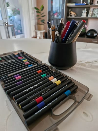 The image shows a collection of black acrylic markers laid out in a transparent case on a white, embroidered tablecloth. The case is positioned in the foreground and is filled with markers, each with a colored band indicating its ink color. Colors visible include orange, pink, yellow, red, blue, green, and purple. In the background, a black cylindrical holder filled with various pens and pencils stands on the same tablecloth. Behind the holder, a shelf is visible with decorative items and a plant. The scene appears to be indoors, likely in a home environment, given the domestic setting of the tablecloth and furniture. There is no indication that this scene is located in Bremen, Germany; no landmarks or identifying features suggest a specific geographic location.