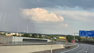 The image captures a highway scene in Hobersdorf, Austria, under a dramatic sky filled with large, white clouds and darker storm clouds. A highway runs through the foreground, curving to the right. To the left of the highway, a noise barrier with a modern, glass-fronted section is visible, followed by a field with several wind turbines. More wind turbines are scattered across the landscape in the distance, along with agricultural fields and some buildings. A blue highway sign indicates directions to "Wien" (Vienna), "Schrick", "Misteltbach-Süd", and "Misteltbach-Ost Wilfersdorf". Several cars and a red truck are on the highway, with no people clearly visible. The overall impression is of a vast, open landscape with industrial infrastructure.