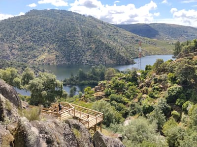 The image depicts the wooden walkways overlooking the Douro River, located in Alijó, Portugal. The scene features a man standing on one of the walkways. In the background, there's a river surrounded by mountains covered in lush greenery and trees. A red and white communications tower stands near the water. The sky is partly cloudy. The foreground consists of rocks and bushes.