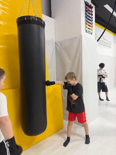 The image displays an indoor martial arts training facility, identified by the signage "Император Школа Единоборств" (Emperor School of Martial Arts), located in Novosaratovka, Russia. The facility features yellow and white padded walls and a light-colored floor.
A black cylindrical punching bag, suspended by chains, hangs against a yellow padded wall. In the center, a young male child with light brown hair, wearing a black t-shirt, red shorts, and black fingerless gloves, is seen striking the punching bag with his right hand. His left hand is in a guard position, and his focus is on the bag.
To the immediate left, partially visible, is another young male facing the punching bag, wearing a white t-shirt and dark shorts. In the background to the right, a third young male with dark curly hair, dressed in a patterned white shirt, dark shorts, and dark socks, is standing and adjusting a black belt around his waist. Mounted on a white wall section in the background is a display of colored martial arts belts, labeled with Russian text including "JUDO" and various principles such as "ВЕРНОСТЬ" (Loyalty) and "ЧЕСТНОСТЬ" (Honesty). A thick black rope also hangs from the ceiling in the background.
