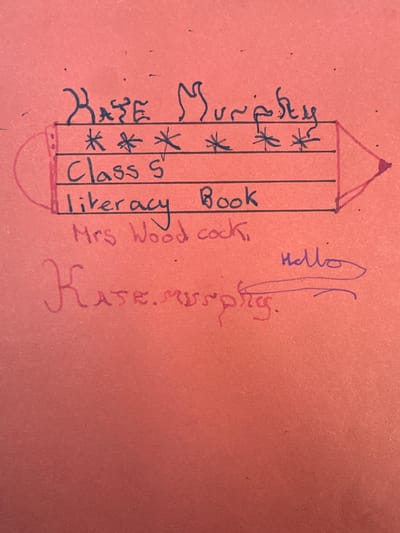 A reddish-orange, textured sheet of paper or thin cardboard is depicted. This item is located in the city of Wealden, United Kingdom. The top portion features text handwritten in black ink. The name "Kate Murphy" is written above a black ink outline of a pencil shape. Within this pencil outline, three horizontal lines contain handwritten text: the first line displays four asterisks ("****"), the second line reads "Class 5", and the third line states "literacy Book". Below the pencil drawing, also in black ink, is "Mrs Woodcock". Further down on the paper, the name "Kate.Murphy." is written in larger, red ink. To the upper right of this red text, the word "Hello" is written in purple ink, accompanied by a wavy purple line extending leftwards from the 'o', partially overlapping the red "Kate.Murphy." text. The paper exhibits minor surface imperfections.