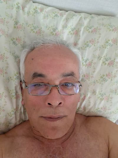 A close-up, over-the-shoulder selfie shows a fair-skinned man with white hair and glasses lying on a floral-patterned pillow. The man's head is tilted slightly to his left, and he is looking directly at the camera. The image is taken indoors, likely in a bedroom. The background consists of the pillow and a white, textured surface, possibly a wall or a headboard. The man's upper chest and shoulder are visible, and his skin appears slightly flushed. The overall lighting is bright and even, suggesting natural daylight. There is no indication of the city of Vidnoye, Russia, in the image itself.