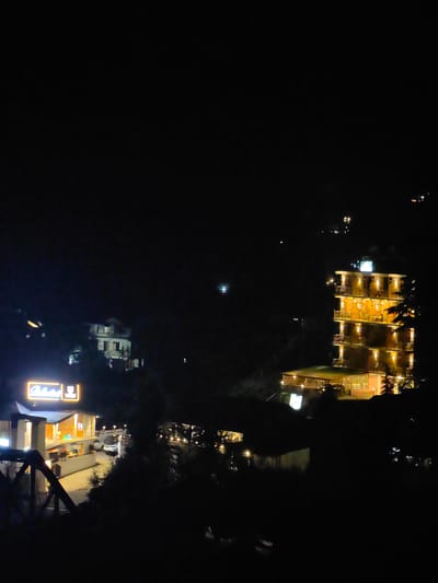 The image shows a nighttime view of a hilly area, possibly in or near the city of Kullu, India. In the foreground, there is a dark, shadowed area that appears to be foliage or a slope. To the left, a modern building with illuminated windows and a glowing sign with text is visible. Further to the right, a multi-story building is lit up, revealing multiple balconies and windows, suggesting it is a hotel or residential complex. The surrounding landscape is dark, with scattered lights from other structures visible in the distance, indicating a developed area within the mountainous terrain. The sky is dark, with a few faint lights that could be stars or distant aircraft.