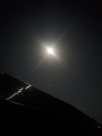 The image shows a night sky with a bright, starburst-like light source, likely the moon or a strong artificial light, positioned in the upper center. Below this, a dark, angled structure, appearing to be a corrugated metal roof, extends from the left side of the frame towards the right. The roof reflects some light, showing a pattern of highlights. The overall scene is dark, with the sky filling most of the frame, suggesting an outdoor setting, possibly in Kason, Nigeria, at night.