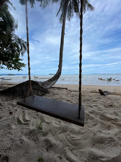 The image depicts a swing hanging from a palm tree on a beach in Baan Sri Thanu, Thailand. The swing is made of wood and is suspended by two ropes. The beach is sandy, and there are some trees and vegetation visible. The sky is blue with some clouds. The water is shallow, and there are some boats in the distance.