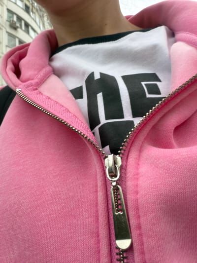 This media file is a close-up, tilted shot of a person wearing a pink zip-up hoodie over a white t-shirt with bold black lettering. The zipper is partially open, revealing the t-shirt underneath. The text on the t-shirt appears to be a stylized logo or word, with thick, blocky letters. The hoodie fabric has a soft, textured appearance, and the zipper pull is a silver metal tab.
The setting appears to be outdoors, with a blurred background suggesting buildings and possibly a street. The image is taken from a low angle, looking upwards towards the person's torso and chest area. The lighting is soft, and there are no strong shadows, suggesting it might be an overcast day. The context provided places the location in Moskovskiy, Russia. No specific activity or event is clearly depicted; it's a still image focusing on the clothing and the textures of the fabric. The overall emotion conveyed is casual and everyday. The visible text on the t-shirt is the most prominent detail besides the clothing itself.