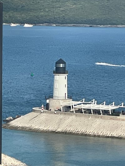 The image depicts a lighthouse situated on a concrete pier extending into a body of blue water. The lighthouse is white with a black lantern room and has several windows. In front of the lighthouse, a white structure with a sloped roof is visible, supported by pillars. The pier is made of concrete blocks and leads to a rocky shoreline. In the distance, a green buoy floats in the water. Several white boats are anchored near a tree-covered coastline in the upper left of the frame. A white jet ski is creating a wake in the water to the right of the lighthouse. The overall environment appears to be a coastal area, likely in the vicinity of Radovići, Montenegro, given the presence of a lighthouse and boats. The lighting suggests it is daytime with clear weather.