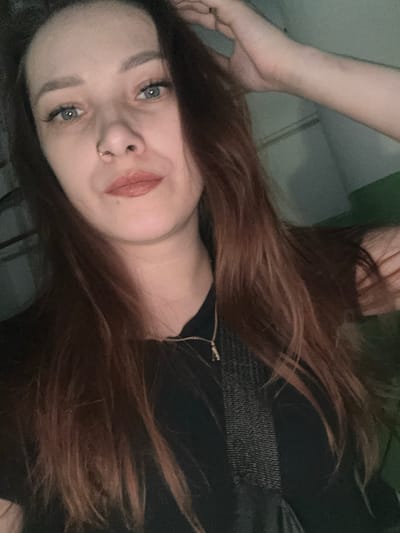 The media file is a close-up, low-angle selfie of a young person lying down, with their head tilted to the side. They have long, reddish-brown hair that falls around their face and shoulders. Their eyes are a striking blue-gray, and they are wearing light makeup, including eyeliner and lip gloss. A small nose ring with a gold hoop is visible. The person is wearing a dark, possibly black, top with a zippered detail. The background appears to be an indoor setting, with hints of a tiled or paneled wall and a dark surface in the foreground. The lighting is dim, creating a soft, somewhat moody atmosphere. The person's expression is neutral to slightly smiling, with a gentle gaze directed at the camera. The overall impression is casual and intimate. No text is visible in the image. The provided location context is Kirov, Russia, but there are no specific visual cues within the image to confirm or deny this. There are no clear indicators of the time of day or weather conditions.