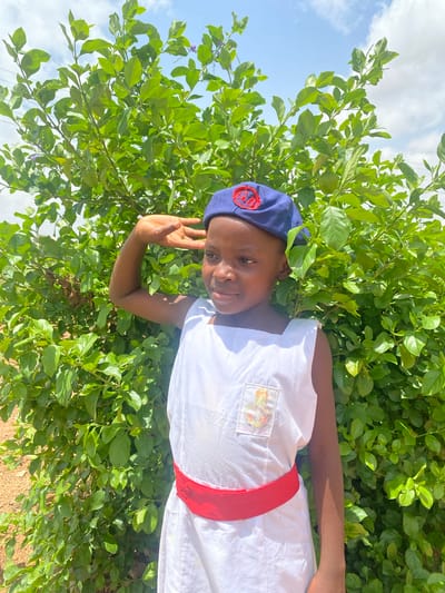 A child in Turu, Nigeria, is photographed outdoors against a backdrop of green foliage and a partially cloudy sky. The child wears a dark blue beret with a circular emblem featuring red and black accents, a white top adorned with a colorful patch on the chest, and a red sash around the waist. The child's right hand is raised, fingers extended in a gesture. The foreground shows sparse vegetation, while the background features lush greenery. The image has a slight tilt, making the vertical axis appear diagonal.