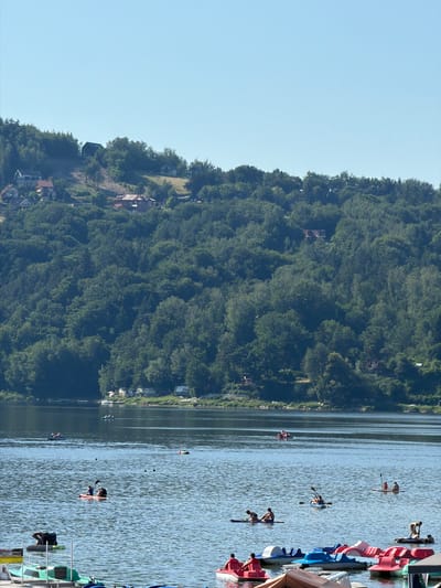 In Gródek nad Dunajcem, Poland, a serene body of water, likely a lake or reservoir, is visible under a clear blue sky. The water reflects the sunlight, creating a shimmering effect. The shoreline is densely populated with lush green trees that ascend a gentle slope. Scattered amongst the trees are several houses, some with red roofs, indicating a settlement on the hillside.

On the water, various individuals are engaged in recreational activities. Several pedal boats are docked or being used near the foreground. Further out, people are seen in kayaks, paddleboards, and other small watercraft, paddling and enjoying the water. Some individuals are sunbathing on paddleboards, while others are actively rowing their boats. A few buoys are dispersed across the water. The overall scene depicts a tranquil summer day at a popular recreational spot.