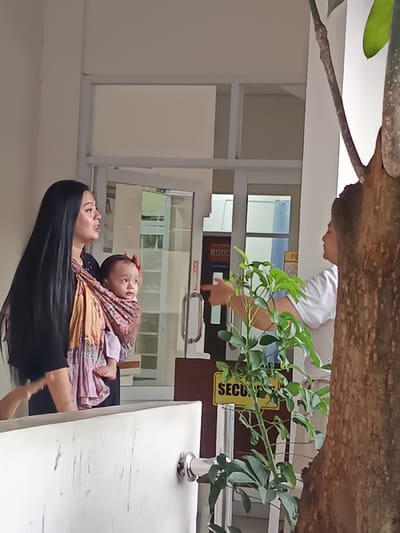 The image displays a scene in Cianjur, Indonesia, within what appears to be a building entrance or a sheltered area. On the left, a female individual with long dark hair, wearing a black top, holds an infant in a patterned fabric baby carrier. The infant, dressed in light clothing and adorned with a red head accessory, faces towards the right. The female's mouth is slightly open, indicating potential speech. To the right, partially obscured by a prominent tree trunk and a green plant, another female individual, wearing a white top, extends her right arm and points with her index finger towards a set of glass double doors centrally located in the background. These doors are equipped with metal handles and lead into an interior space. A yellow sign with the partial text "SECURI" (likely "SECURITY") is visible on a brown counter positioned in front of the doors. A white barrier or railing occupies the lower-left foreground. The scene depicts an interaction between the two adult females, one carrying an infant, at a building's entry point.