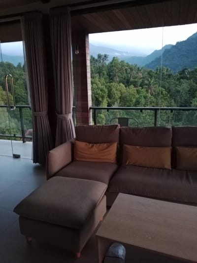 The image depicts a covered patio or balcony area with a sectional sofa and an ottoman. The furniture is upholstered in a neutral fabric, with orange accent pillows. A low wooden coffee table is positioned in front of the sofa.

Through a large glass door and window, a lush, green landscape is visible. This includes dense tropical foliage, palm trees, and rolling hills or mountains in the distance. The sky appears overcast or misty, suggesting it may be early morning or late evening.

The setting appears to be a residential or hospitality location, likely designed to offer expansive views of the natural environment. Given the tropical vegetation and mountainous terrain, the location is identified as Sawan, Indonesia. The scene is peaceful and suggests a comfortable, relaxing atmosphere.