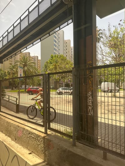 The image shows a street scene in L'Hospitalet de Llobregat, Spain, likely during the daytime. In the foreground, a dark metal fence runs horizontally across the frame, with a concrete curb below it. A person is seen riding a bicycle with a neon green frame through an opening in the fence. The cyclist is partially visible, wearing shorts and a t-shirt, with their back to the camera. Behind the fence, a paved area is visible, with a white van and a red car parked. Several tall, modern buildings and palm trees are in the background, suggesting an urban environment. A sign with text, possibly a traffic or pedestrian sign, is also visible near the fence. The overall impression is of a sunny day in a residential or mixed-use urban area.