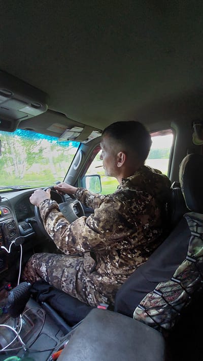 An exterior, eye-level, medium shot captures a man wearing camouflage clothing driving a car on a paved road surrounded by greenery. He is holding the steering wheel with both hands. The car's interior is visible, including the dashboard, windshield, and side mirror. The man's face is turned to the right, and he appears to be looking in the side mirror. The environment outside the car is a blur of trees and foliage, suggesting movement. The image is taken from within the car, looking towards the driver. The lighting suggests it is daytime, with overcast conditions. The image provides no specific clues about the exact location of Severnye Koryaki, Russia, beyond the general context of a natural, rural environment.