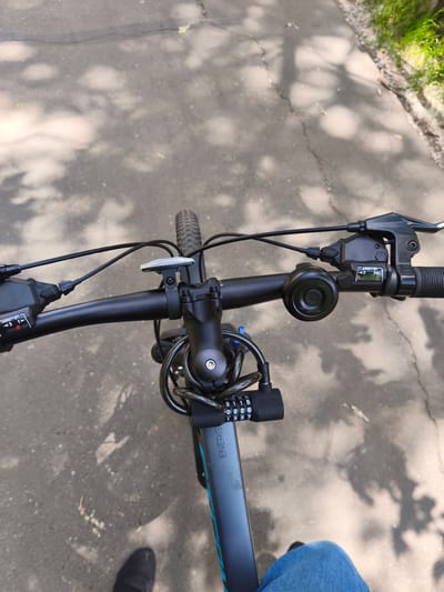 The image is a first-person perspective of someone sitting on a bicycle, looking down at the handlebars and the road ahead. The scene appears to be an outdoor location, likely a park or a paved path, as evidenced by the asphalt surface and the dappled sunlight filtering through tree leaves. The bicycle is black, with visible brake levers and gear shifters on the handlebars. A black combination lock is attached to the frame of the bicycle, with the numbers 9115 and 0226 visible. The word "ENERGY" is printed vertically on the bicycle's frame. The rider is wearing blue jeans. The environment suggests a daytime setting with shadows cast by trees on the ground. No specific landmarks of Komsomolsk-on-Amur, Russia, are identifiable in the image.