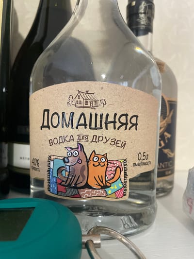 The image shows a close-up of a clear glass bottle of vodka with a kraft paper label. The label features the Russian text "ДОМАШНЯЯ ВОДКА ДЛЯ ДРУЗЕЙ", which translates to "Homemade Vodka for Friends". An illustration on the label depicts an orange cat and a brown dog sitting on a patterned rug. The label also indicates "0.5L" (capacity) and "40%" (alcohol content). The bottle is surrounded by other bottles and a green object. The location is not apparent from the image.