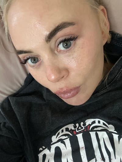 A fair-skinned person with light blonde hair is positioned on a soft surface, likely a bed or pillow, wearing a dark gray or black t-shirt with a white graphic design. The person's face is the focal point, featuring prominent eyebrows, extended eyelashes, and light-colored eyes. The skin appears smooth and possibly slightly dewy, with a natural lip color. The image does not reveal any information about the city of Высокий, Russia. The scene appears to be indoors, well-lit, and the focus is primarily on the person's facial features.