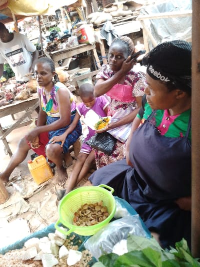 In an outdoor market, three women and a child are pictured. The leftmost woman sits on a surface near a yellow container and a table displaying meat. The child sits in the center holding a styrofoam container of food. A woman wearing a pink-trimmed dress is adjusting something behind her ear, and another woman wearing a "Reebok" hat and a blue apron sits on the right. A green basket of seeds is in the foreground.