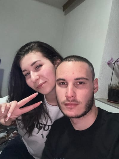 The image depicts two young adults, one female and one male, in an indoor setting within Erma Reka, Bulgaria. The female, positioned on the left, has long dark hair and a light complexion, looking towards the camera with a slight smile. She is wearing a white t-shirt with "GUES" visible on the front and displays a peace sign with her left hand, which has light-colored nails with faint heart designs. The male, positioned in the foreground on the right, has short buzzed hair and a light beard, looking directly at the camera with a neutral expression. He is wearing a black t-shirt. The background consists of a plain, light-colored wall. A wooden beam or molding is visible at the top left corner of the frame. In the upper right background, a potted plant with purple flowers rests on a white shelf or ledge. The scene is illuminated by frontal, artificial light.