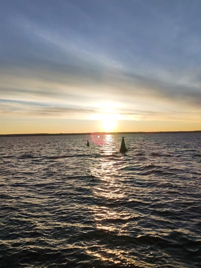 The image shows a body of water, likely a lake or bay, at sunset. The sun is setting on the horizon, casting a bright glow and reflecting off the water's surface. There are two navigation buoys visible in the water. The sky is a mix of blue and orange hues, with clouds scattered across the horizon. The location may be in the vicinity of Myaksa, Russia. The scene does not involve any visible people or explicit actions. The environment is characterized by the tranquility of the water and the soft light of the sunset.