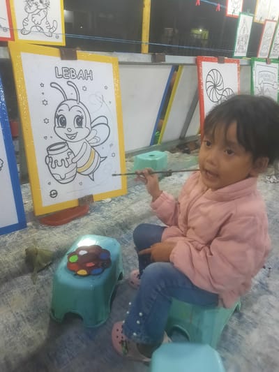A young child, presumably female, with dark hair, is depicted in a close-up, slightly elevated shot, engaged in painting at an outdoor or semi-outdoor venue in Sukasari, Indonesia. The child wears a light pink zippered puffer jacket, blue jeans with pearl-like embellishments on the left leg, and pink Crocs-style shoes. She is seated on a small, light blue plastic stool. Her right hand holds a slender paintbrush, directed towards a coloring page displayed on a yellow-framed easel in front of her. The coloring page features a line drawing of a cartoon bee ("LEBAH" in Indonesian) holding a pot of honey, with some yellow color already applied to the bee's body. Scattered stars and hearts are also drawn on the page.

In the immediate foreground, another identical light blue plastic stool holds a brown, flower-shaped paint palette containing at least eight distinct colors: red, green, blue, yellow, white, brown, black, and orange. A dried, light brown leaf rests on the patterned gray ground covering to the left of the stool.

The background reveals a setup with multiple similar easels and coloring pages. To the left, a blue-framed easel is partially visible. To the right, several finished or in-progress coloring pages are hung on a string with clothespins; one red-framed page displays a concentric spiral design, and another green-framed page reads "Marhaban Ya Ramadhan." The overall environment is dimly lit, indicative of nighttime, with a distant structure showing faint illumination.