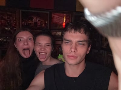 The image shows an upside-down selfie of three young adults, two women and one man, in what appears to be a dimly lit establishment. The subjects are close together, with their faces towards the camera, suggesting a playful or informal gathering.
The man on the left has light blue eyes and dark hair. He is wearing a black t-shirt. One of the women, in the center, has her tongue sticking out, her mouth open in a wide smile, and appears to be wearing a grey t-shirt. The woman on the right also has her mouth open, showing her teeth and tongue, and her eyes are wide. She has long, dark, wavy hair.
The background is dark and appears to be a wall adorned with several framed pictures or posters. Some of the frames are dark wood, and the items within them are not clearly discernible due to the low light and the upside-down orientation. There are reflections in what might be glass covering these frames, showing some interior lights and possibly other patrons or decor. A neon red light source is visible in one of the frames, suggesting the establishment could be a bar or a restaurant with ambient lighting.
The overall mood of the image is energetic and fun, conveyed by the exaggerated facial expressions and close proximity of the individuals. The time of day is likely evening or night, given the dim lighting. The location cues are not specific enough to pinpoint an exact place within Jing'an District, China, beyond it being an indoor venue. No visible text can be clearly identified.