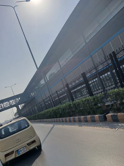 A light yellow, four-door hatchback with a license plate that reads "AXK 423 ICT-Islamabad" is driving on a multi-lane road. The car is angled to the right, showing its rear passenger side. In the background, a modern, elevated structure with a corrugated metal roof and a blue accent line runs parallel to the road. This structure appears to be a transit station or overhead walkway. A pedestrian bridge connects to this elevated structure from the left. The area is lined with manicured green bushes and a curb marked with alternating black and yellow stripes. The sky is bright and hazy, suggesting a sunny day. The scene is set in Peshawar City Tehsil, Pakistan.
