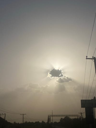 The image depicts a bright, hazy sky, likely taken in Unguwar Fatika, Nigeria, though this is not explicitly visible in the image. The sun is obscured by a dark cloud, creating a dramatic effect with rays of light emanating outward. In the upper part of the frame, there are utility poles and power lines. On the right edge of the frame, there is some signage and what appears to be a metal structure, and a dark area of trees.