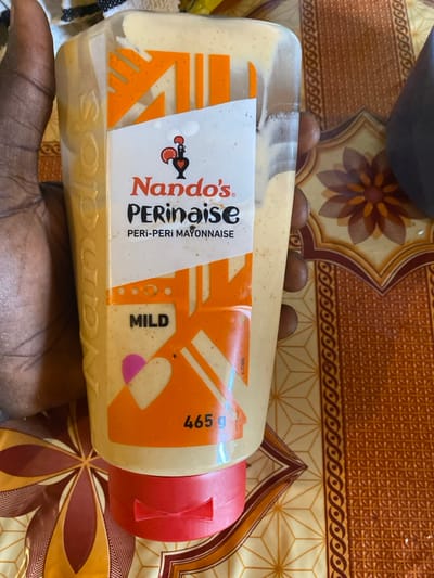 A bottle of Nando's Perinaise is shown horizontally, held from below by a dark-skinned hand, against a patterned tablecloth or mat. The background features intricate floral and geometric designs in shades of brown, orange, and beige. Three small, round, brown objects, possibly seeds or nuts, are visible behind the top edge of the bottle.

The bottle's label clearly displays the brand "Nando's" with its distinctive rooster logo, followed by "PERi-PERi MAYONNAISE". A large "MILD" indicator is prominent on the orange-colored part of the label, along with "465 g" indicating the net weight. The sauce inside the bottle appears creamy and light yellow. The bottle is capped with a bright red lid. The scene is set indoors, likely on a table in Lakka, Sierra Leone.