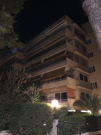 The image captures a multi-story building, likely an apartment complex, at night in Marousi, Greece. The building occupies the left and central parts of the frame, with lush vegetation and a prominent street light on the right.
The building is constructed from a light-colored material, possibly beige or pale yellow, and features numerous floors. Each floor has a balcony with metal railings. Several balconies are equipped with brown or beige retractable awnings, and some show small plants or greenery. Windows across the facade are a mix of dark reflections and those revealing interior lights. Two satellite dishes are visible on one of the lower-level balconies.
The right side of the image is dominated by dense green foliage, including large bushes, a tall palm tree, and other leafy plants in the foreground. A bright street light, emitting a warm yellow-white glow, stands on the right, illuminating the surrounding vegetation and parts of the building.
The scene is set at night, indicated by the dark sky, the artificial lighting from the street lamp and building interiors, and the overall low-light conditions. No people are visible. A small blue sign with indistinct white markings is attached to the pole of the street light. The atmosphere appears calm.
